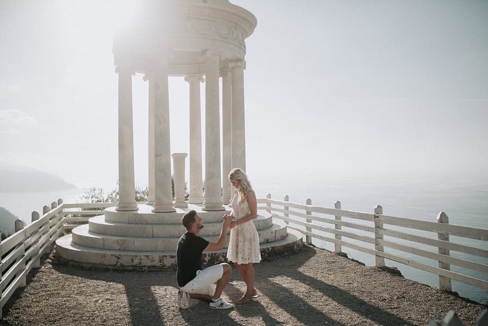 Verlobungsantrag am Meer vor einer weißen Säule mit Blick auf Wasser und Berge.