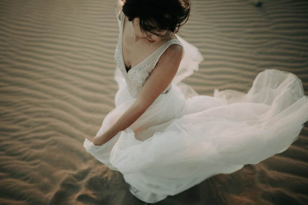 Elegante Braut im weißen Kleid in Sand mit windigem Rock, kreative Hochzeitsfotografie, emotionale Atmosphäre, Natur inspired.