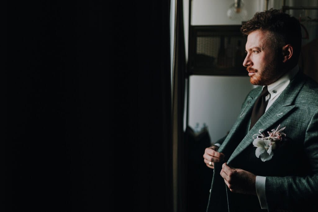 Bräutigam im grünen Anzug mit Blume am Revers, blickt aus dem Fenster. Hochzeitstag.