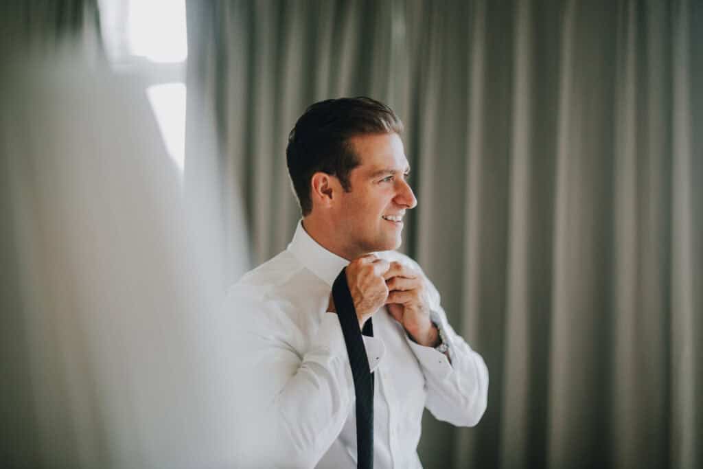 Bräutigam bindet Krawatte. Vorbereitung auf Hochzeit in Irland. Elegantes Outfit, helles Interieur.