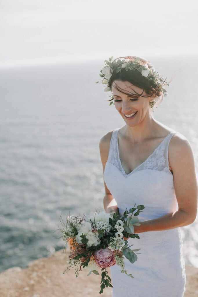 Braut mit Blumenkranz und Brautstrauß am Meer, lächelnd. Hochzeitsfotograf Irland.