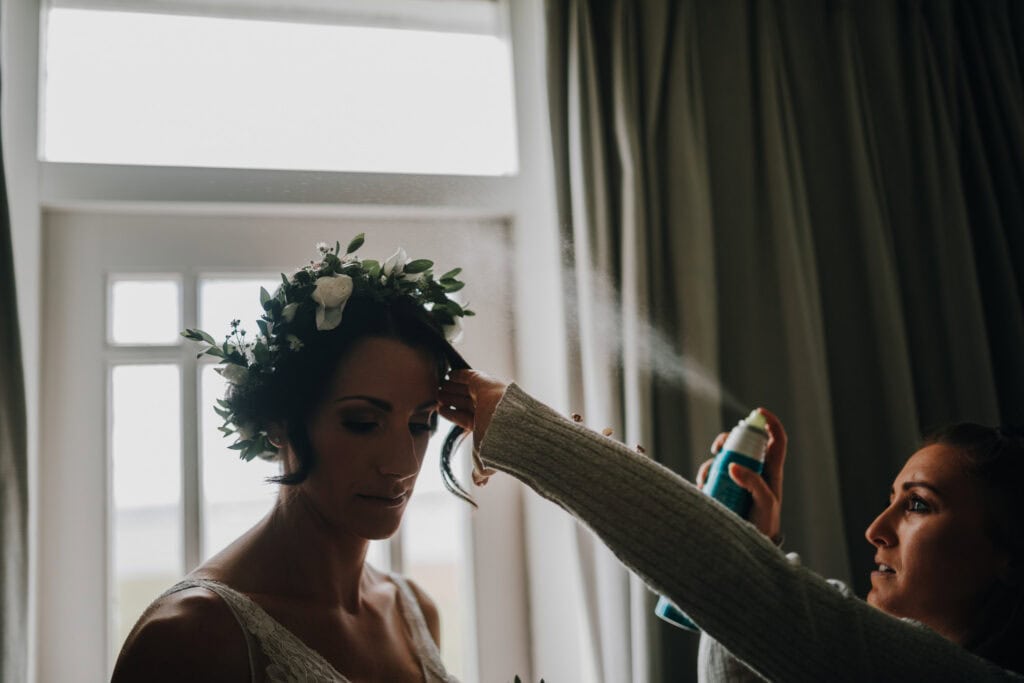 Brautstyling mit Blumenkranz: Friseur sprüht Haarspray vor Fenster. 