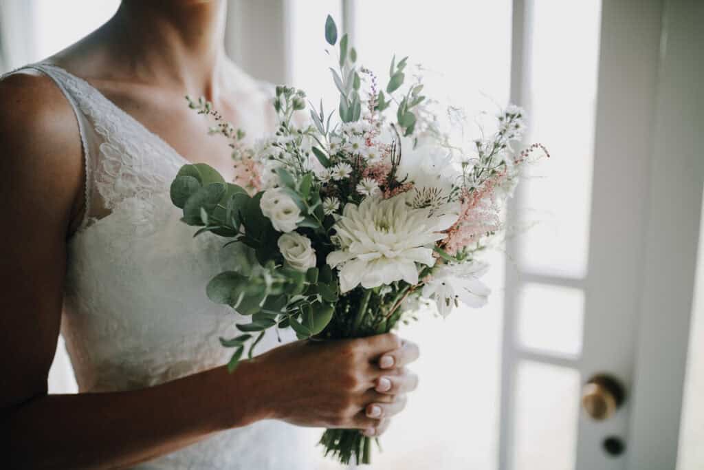 Braut hält weißen Blumenstrauß mit Eukalyptus vor weißem Kleid. Zarte Hochzeitsdetails.