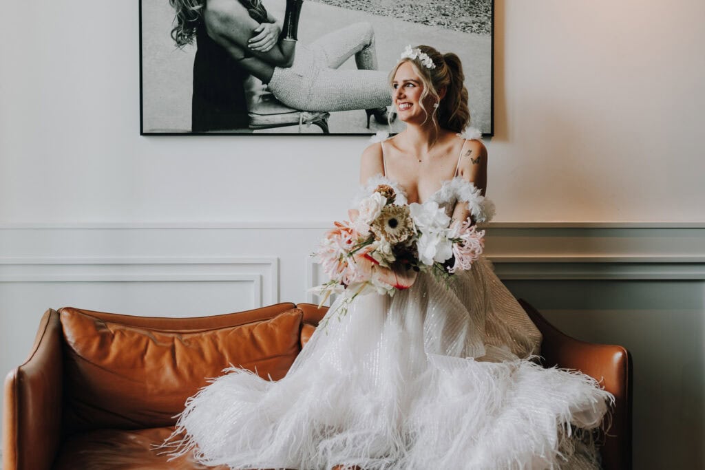 Braut mit Blumenstrauß sitzt auf Ledercouch, Hochzeitsfotograf München.