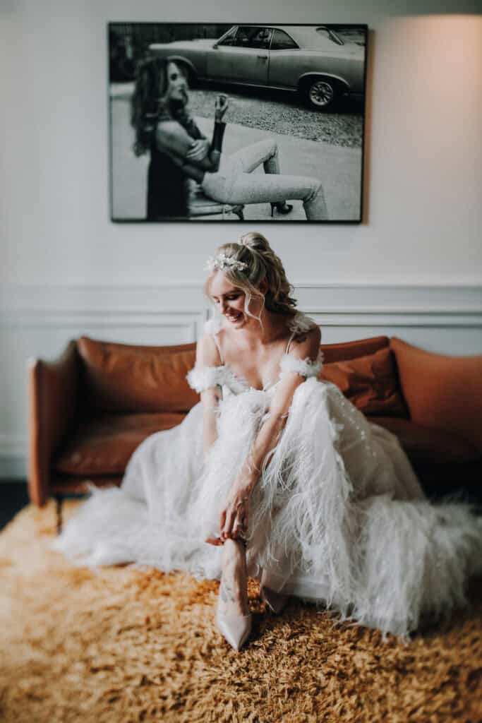 Braut im Federkleid sitzt auf Sofa, lächelt. Detailaufnahme der Hochzeitsfrisur und des Kleides.