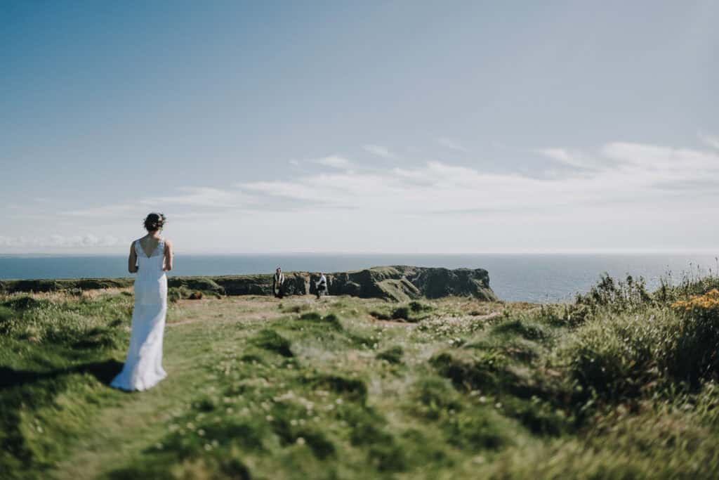 Braut in Irland an Klippen, Hochzeitsfotograf im Hintergrund. Weites Meer und blauer Himmel.