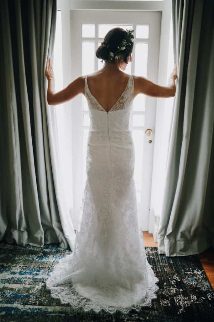 Braut mit Blumenschmuck im Haar vor Fenster mit Vorhängen. Rückenansicht des Brautkleides.