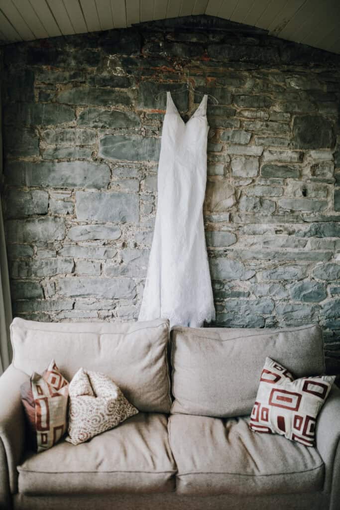 Weißes Brautkleid hängt vor einer Steinmauer über einem beigen Sofa mit gemusterten Kissen.