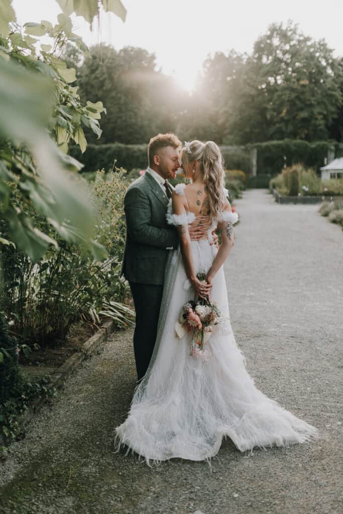 Brautpaar umarmt sich im Garten, die Braut hält einen Blumenstrauß. Sonnenlicht im Hintergrund.