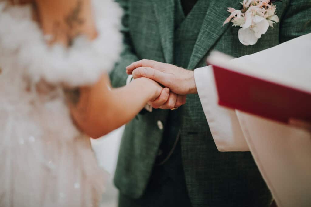 Brautpaar hält Hände bei Trauung, Bräutigam im grünen Anzug mit Blumenanstecker. München Hochzeitsfotograf.
