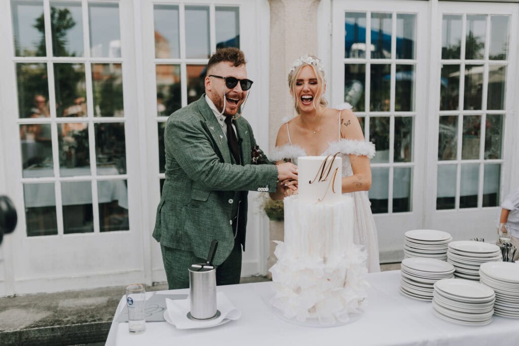 Brautpaar schneidet Hochzeitstorte an. Glückliche Stimmung, festliche Kleidung, Location im Freien.