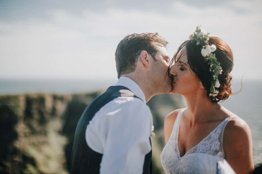 Brautpaar küsst sich innig vor irischer Küstenlandschaft. Braut mit Blumenkranz. Hochzeitsfotograf Irland.