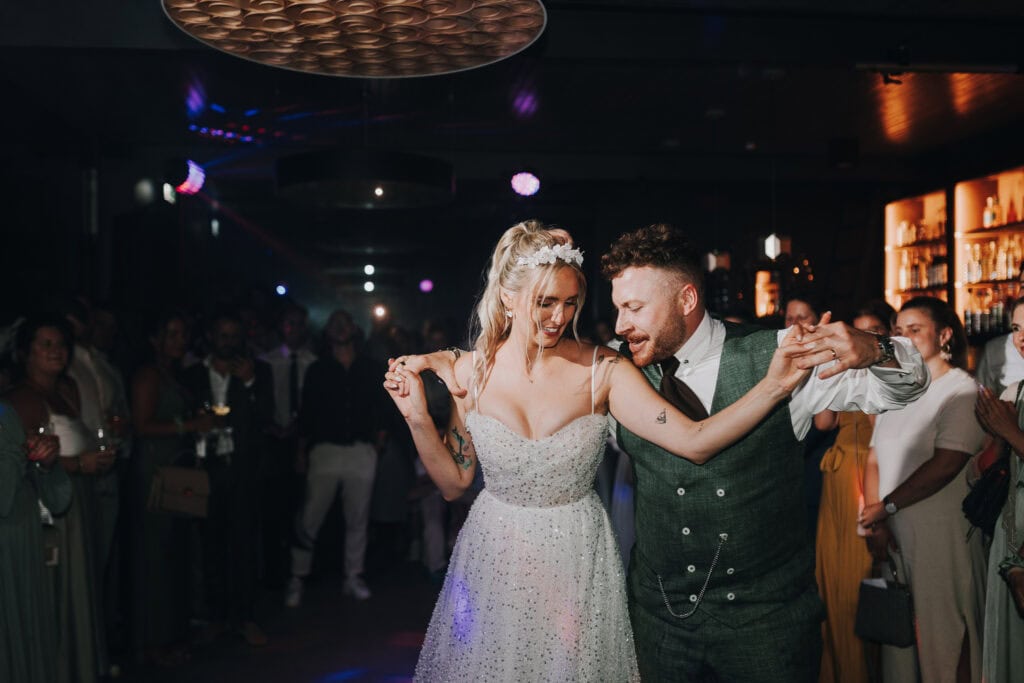 Brautpaar tanzt auf Hochzeit in München. Braut im glitzernden Kleid, Bräutigam in Weste. Fröhliche Hochzeitsgesellschaft.