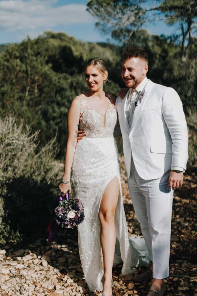 Brautpaar in Weiß posiert vor grüner Landschaft.