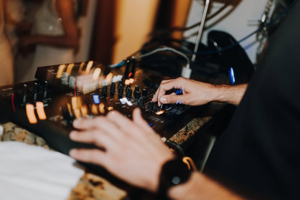 DJ-Pult in Aktion, Hände bedienen Mischpult. Event oder Hochzeit im Hintergrund unscharf.