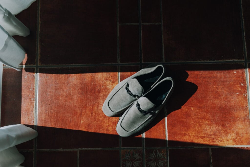 Graue Wildlederschuhe auf rotem Fliesenboden mit Sonnenlicht und weißen Stoffen. 