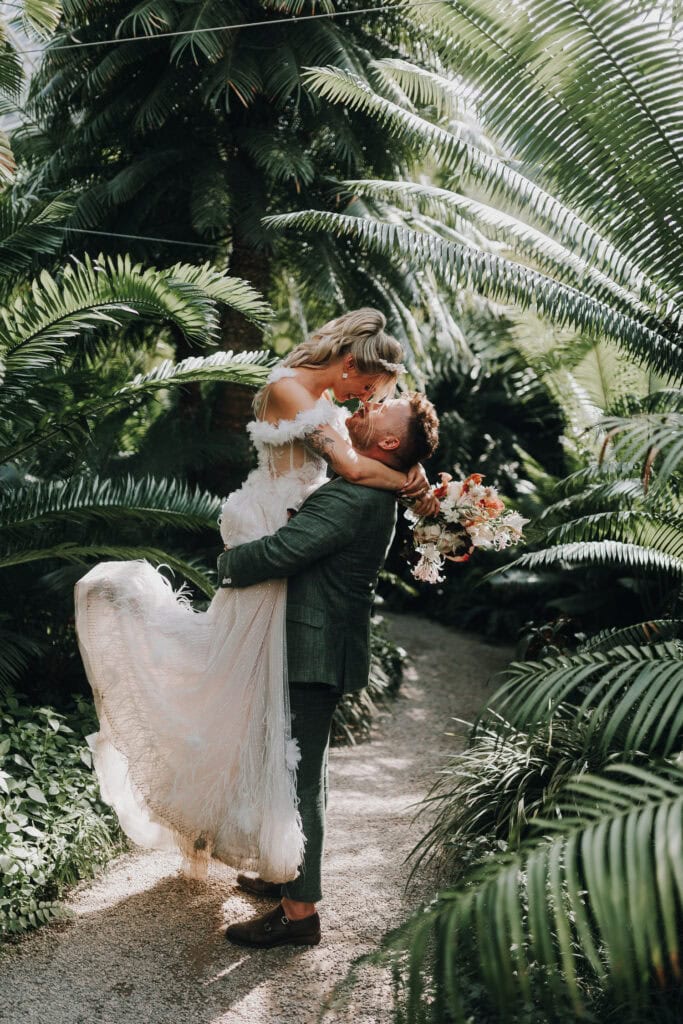 Bräutigam hebt Braut im Brautkleid mit Blumenstrauß in üppigem Gewächshaus hoch. Hochzeit München.