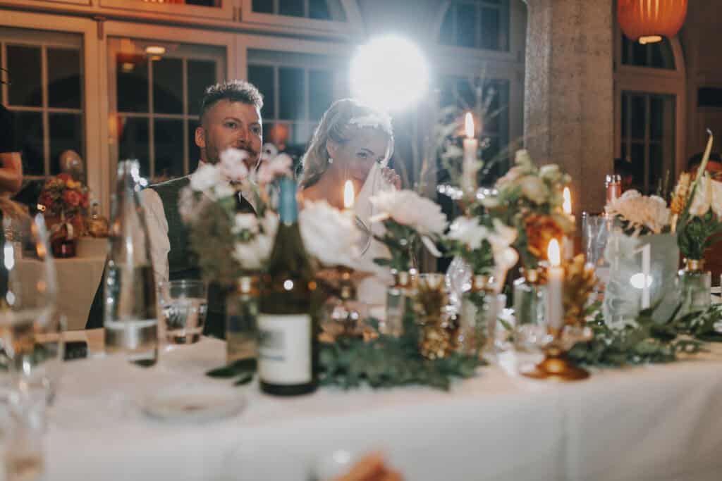 Braut weint gerührt bei Hochzeitsrede, Bräutigam schaut zu. Kerzen und Blumen schmücken den Tisch.