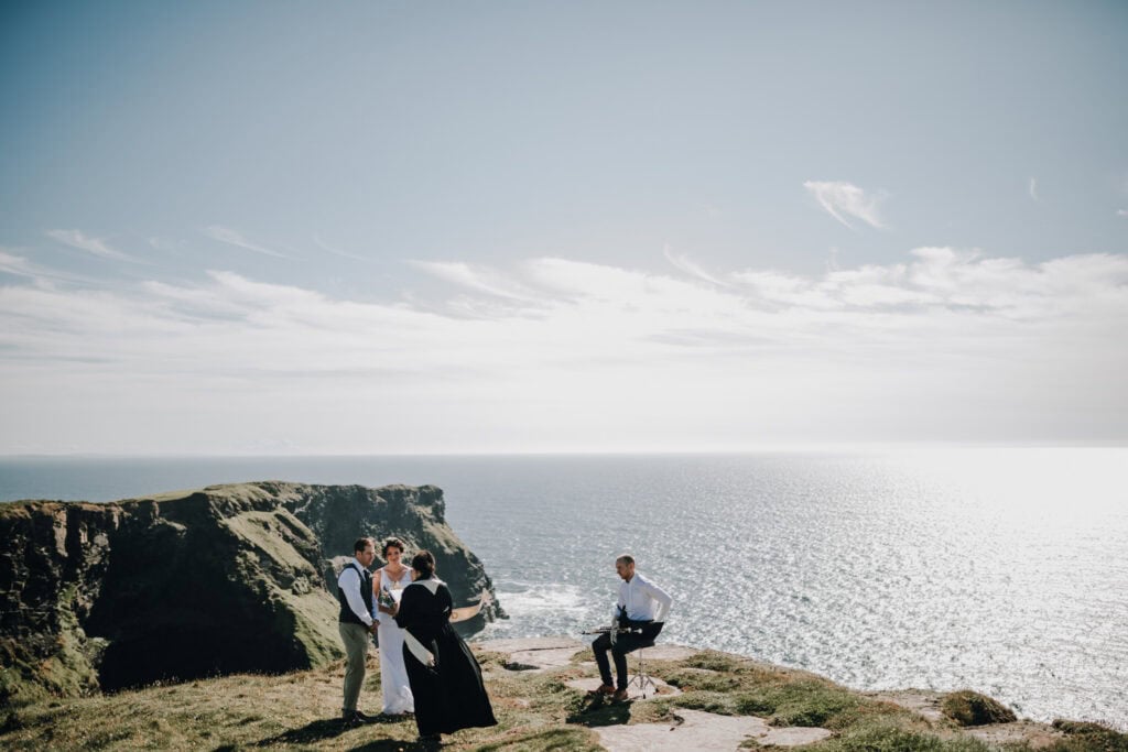 Hochzeit an den Cliffs of Moher, Irland. Brautpaar mit Redner und Musiker vor atemberaubender Küstenlandschaft.