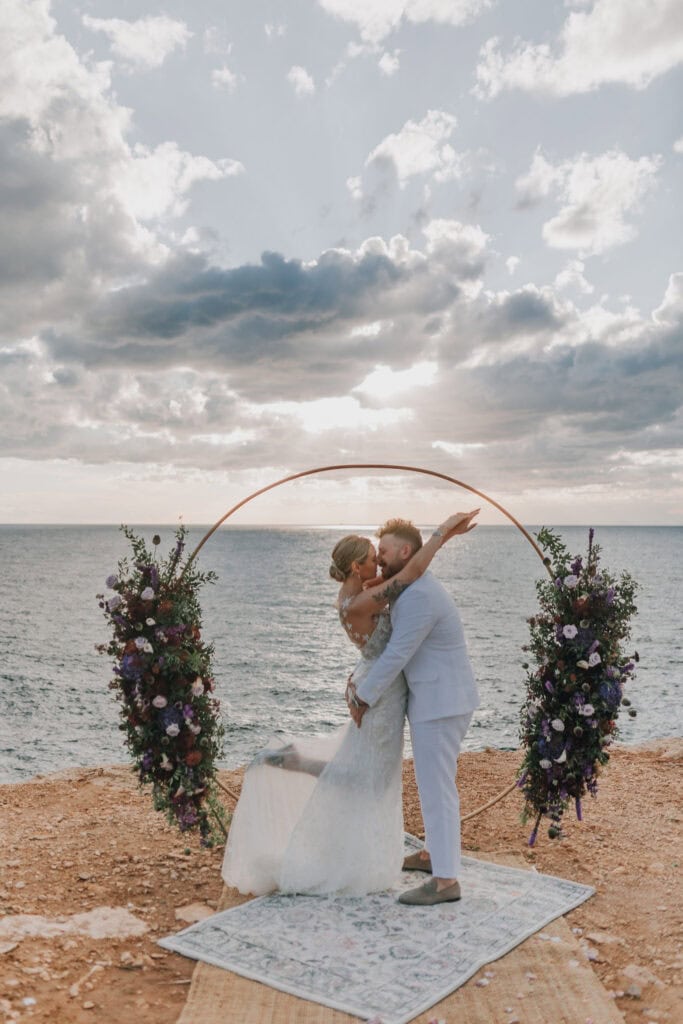 Brautpaar küsst sich vor einem Blumenbogen am Meer. Ibiza Hochzeitsfotograf. Sonnenuntergang.