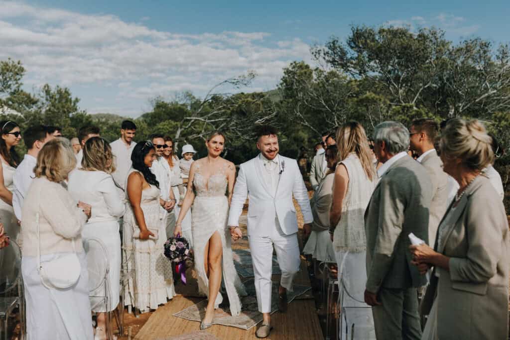 Brautpaar in Weiß schreitet durch Hochzeitsgäste auf Ibiza. Boho-Stil, sonniger Tag.