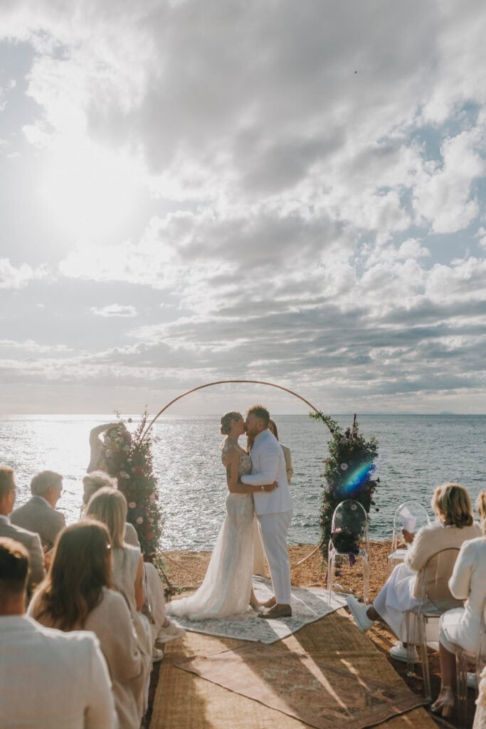 Brautpaar küsst sich vor Bogen am Meer. 