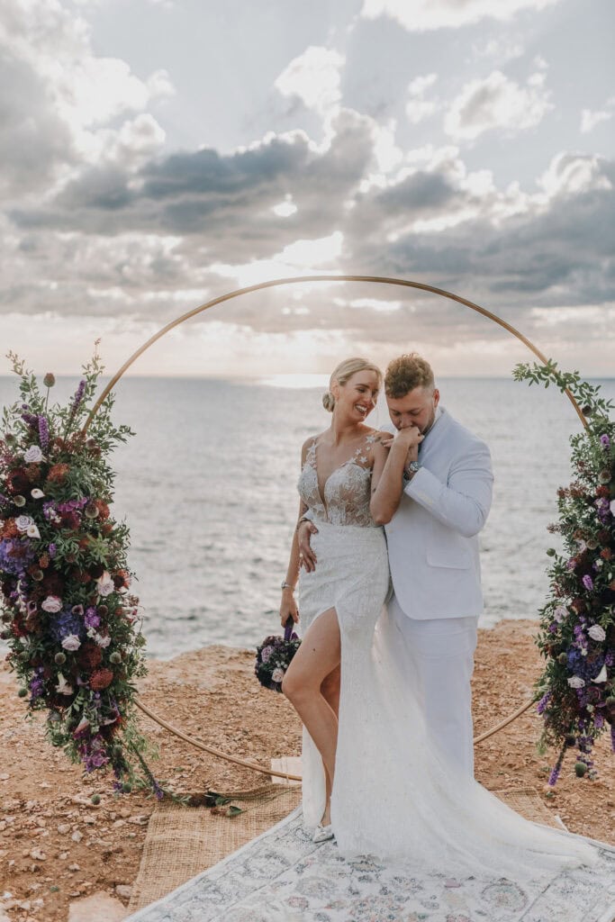 Brautpaar umarmt sich vor dem Meer auf Ibiza. Goldener Hochzeitsbogen mit Blumen. Hochzeitsfotograf Ibiza.