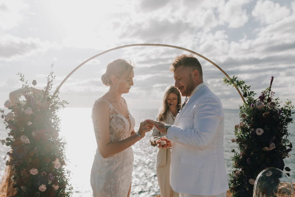 Brautpaar tauscht Ringe bei Hochzeit auf Ibiza unter goldenem Bogen mit Blumen.