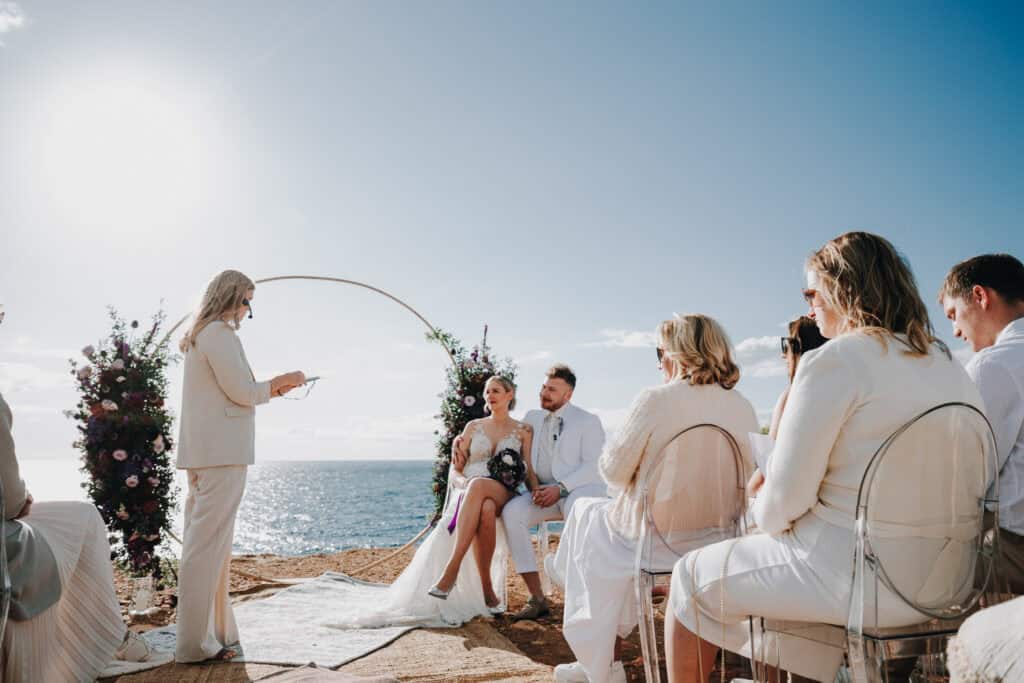 Brautpaar sitzt bei Hochzeit auf Ibiza vor Meer. Traurednerin und Gäste in Weiß.