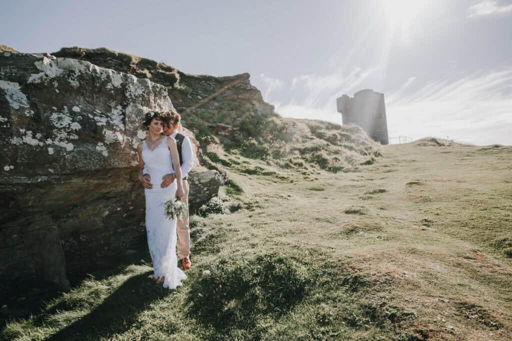 Brautpaar umarmt sich vor irischer Landschaft mit Turm. Blumenkranz und Brautstrauß.