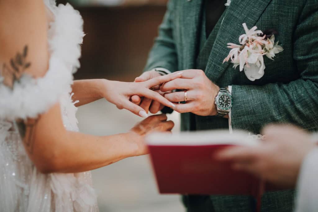 Bräutigam steckt Braut Ring an. Detailaufnahme von Händen bei der Trauung. Blumenanstecker, Uhr sichtbar.