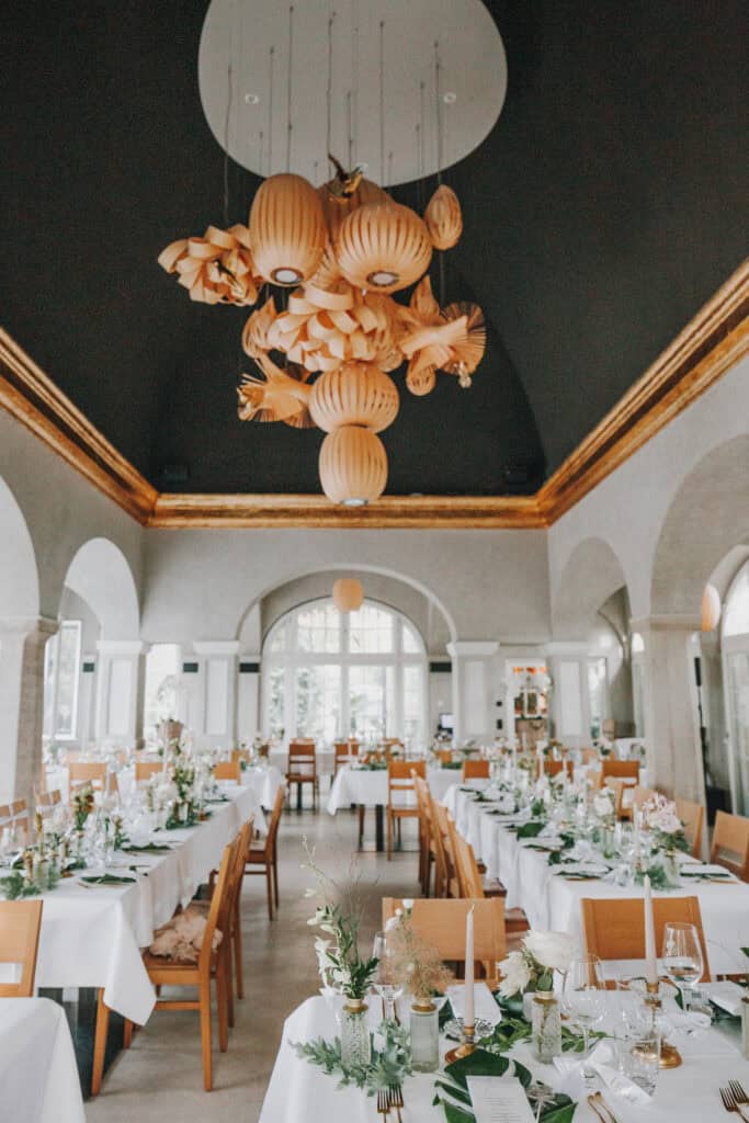Elegante Hochzeitsdekoration mit langen Tischen, Blumenarrangements und einer kunstvollen Deckenleuchte.