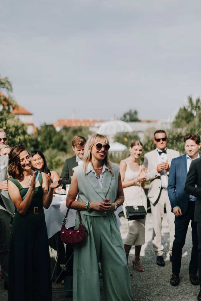 Hochzeitsgäste applaudieren bei einer Hochzeit im Freien, stilvolle Kleidung, sonniger Tag.