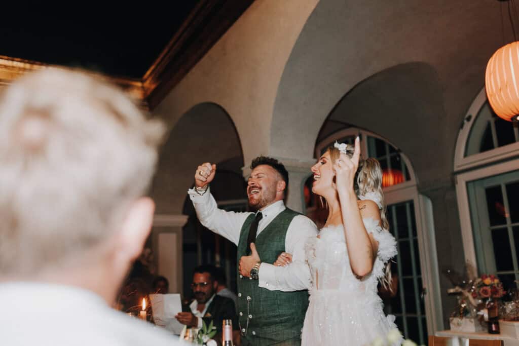 Fröhliches Hochzeitspaar jubelt mit Gästen. Braut im weißen Kleid, Bräutigam im grünen Anzug. München Hochzeitsfotograf.