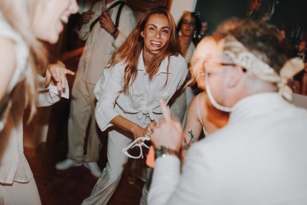 Fröhliche Frau tanzt auf Hochzeitsparty in Ibiza, gekleidet in Weiß. Lachende Gäste im Hintergrund.