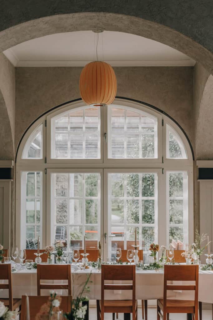 Hochzeitstafel mit Blumenschmuck unter einem Bogenfenster. Warme Farben und einladende Atmosphäre. Hochzeitsfotograf München.