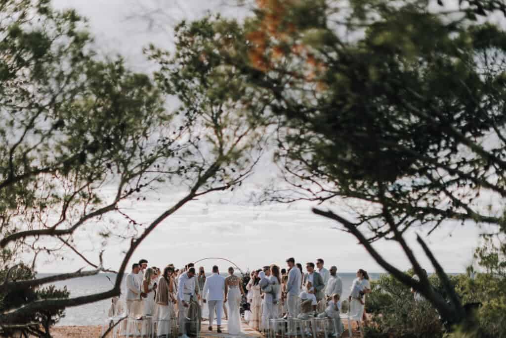 Hochzeitszeremonie im Freien auf Ibiza mit Brautpaar, Gästen und Meerblick unter Bäumen.