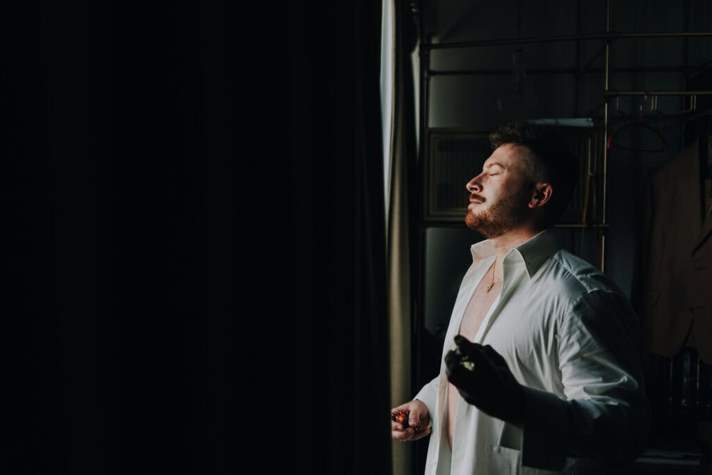 Mann mit Bart öffnet weißes Hemd im goldenen Licht, Kette sichtbar.